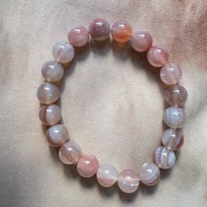 Könnte beinhalten: Armband mit runden Perlen in Rosa-, Weiß- und Pfirsichtönen. Die Perlen haben ein marmoriertes Aussehen. Das Armband ist elastisch und zum Tragen am Handgelenk konzipiert. Die Perlen haben einen Durchmesser von ca. 1 cm.