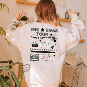 May include: A white sweatshirt with a black graphic design featuring the text "The Eras Tour" and various black and white illustrations, including a clapperboard, a ticket stub, a microphone, a guitar, a fan, and a camera. The text "Surprise Songs" is also included in the design.