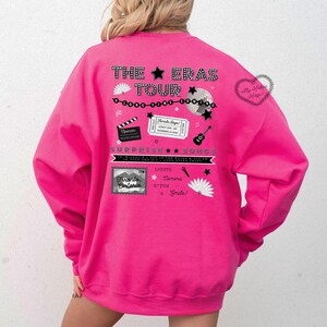 May include: Pink sweatshirt with a graphic design featuring the text "The Eras Tour" and various images including a star, a microphone, a camera, a guitar, and a ticket stub. The text "Surprise Songs" is also included.