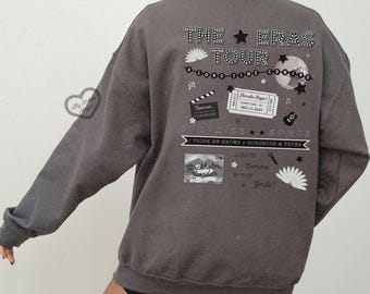 Puede incluir: Una sudadera gris oscuro con un diseño gráfico en la espalda. El diseño incluye el texto "THE ERAS TOUR" y varias ilustraciones como una bola de discoteca, estrellas y un boleto. El texto adicional incluye "SURPRISE SONGS" y "Stockholm, Sweden."