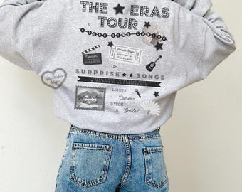 Puede incluir: Una sudadera gris con el texto "THE ERAS TOUR" y varios gráficos, incluyendo estrellas, un boleto y un micrófono. La parte trasera de la sudadera presenta texto y diseños adicionales, creando un look único y llamativo.