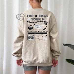 May include: A beige sweatshirt with a black graphic print on the back. The graphic features the text "The Eras Tour" with stars, musical notes, and other design elements. The text "Surprise Songs" is also included in the graphic.