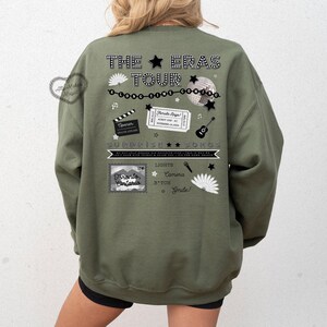 May include: An olive green sweatshirt with a graphic design on the back. The design includes text that reads "THE ERAS TOUR" with various icons such as a film clapper, ticket, guitar, and a stamp that says "Toronto, Ontario".