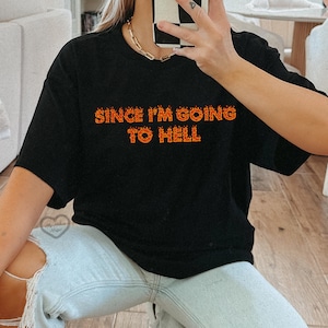 Pode incluir: Camiseta preta com o texto "Since I'm Going To Hell" em laranja com chamas ao redor das letras.