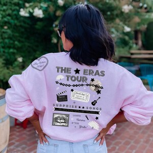 May include: Pink sweatshirt with a graphic design featuring the text "The Eras Tour" with a star and other decorative elements. The text "Surprise Songs" is also included with a list of song titles: "Lights", "Camera", "B*tch", and "Smile!"