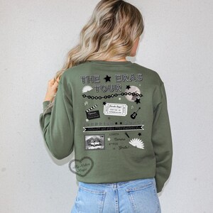 May include: Olive green sweatshirt with a graphic design on the back. The design includes the text "THE ERAS TOUR" with stars, a disco ball, a film clapper, a ticket, a guitar, and other decorative elements. The text "Toronto, Ontario" is also visible.