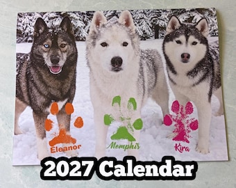 May include: A 2027 calendar featuring three husky dogs in a snowy landscape. Below each dog is a colored paw print with a name: orange for Eleanor, green for Memphis, and pink for Kira. The text 2027 Calendar is printed at the bottom.