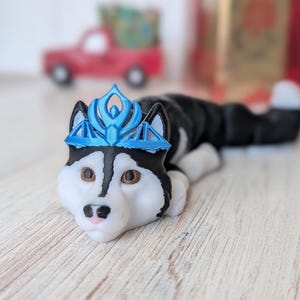 May include: A figurine of a husky dog wearing a blue crown. The dog is black and white with brown eyes and a pink nose. The figurine is lying down on a wooden surface. A red toy truck is in the background.