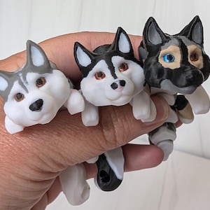 May include: Three small articulated husky dog figurines in grey, black, and white. The left figure is grey and white, the center is black and white with brown eyes, and the right figure is black and grey with one blue eye and one brown eye.