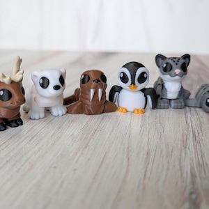 May include: A set of six small animal figurines in a cartoon style. The collection features a brown reindeer, a white polar bear, a brown walrus, a penguin, a grey leopard, and a grey rhinoceros. Each figure has large, black eyes.