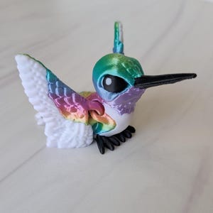 May include: A colorful hummingbird figurine with a white body, rainbow wings, and a black beak. The bird's head is a gradient of green, blue, and purple. The wings are partially spread, and the feet are black.
