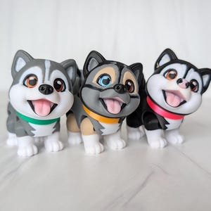 May include: Three cartoon husky dog figurines. The dogs have gray, black, and white fur with brown eyes and pink tongues. Each dog wears a collar in green, orange, and pink. The figurines are smiling and standing.