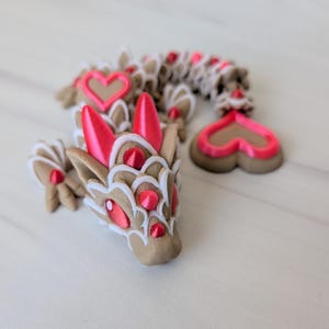 May include: A decorative dragon figurine with a tan body, red accents, and white trim. The dragon features heart-shaped details and red gem-like eyes. The figurine is lying on a white surface.
