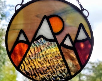 May include: A stained glass suncatcher depicting a mountain range with a setting sun. The mountains are a mix of brown, orange, red, and white glass. The sun is a bright orange.