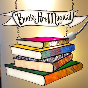 Puede incluir: Una ventana de vitral que cuelga con una pancarta que dice "Books Are Magical". La pancarta está suspendida de dos cadenas. Debajo de la pancarta hay una pila de cinco libros de vitral con diferentes vidrios de colores. Los libros están iluminados desde atrás, creando un brillo cálido.