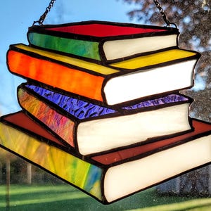 Puede incluir: Vidriera que representa una pila de libros con cubiertas coloridas. Las cubiertas son rojas, naranjas, verdes, amarillas y azules. Las páginas son blancas y los bordes negros. Una cadena está adjunta para colgar.