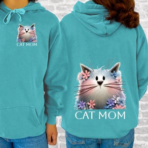 May include: Teal hoodie featuring a cat graphic with floral accents. The front and back of the hoodie display the same design: a fluffy cat face surrounded by flowers, with the text "CAT MOM" below the image. The hoodie has a drawstring hood.