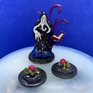May include: Miniature fantasy figure of a robed character with a hood, holding a book, and accompanied by purple tentacles. Two small, round, dark platforms with pink and green objects are in front of the figure.