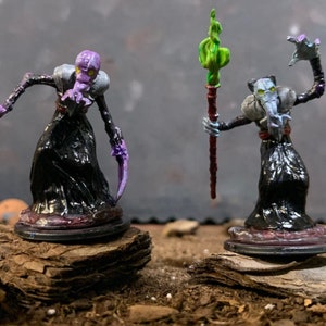 May include: Two painted miniature figures of humanoid creatures with long, thin limbs and large, bulbous heads. The figures are wearing black robes and have purple and green accents. One figure is holding a staff with a green flame at the top.