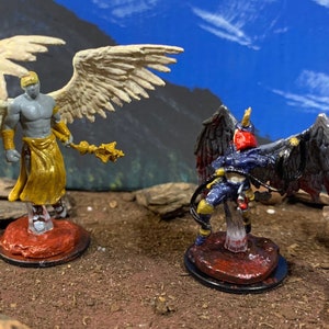 May include: Two miniature figurines of winged characters. One figure is a gray-skinned angel with gold accents and a staff. The other is a dark-winged figure in blue and red armor. Both figures are on red bases, set against a backdrop of a blue sky.