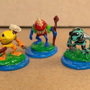 May include: Three miniature fantasy frog figurines. One is yellow with orange spots and a chef's hat, another is red and blue with a horn, and the third is green and black with a silver shoulder plate. Each stands on a green base surrounded by blue.