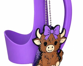 May include: A purple silicone cup holder handle featuring a dangling charm of a brown highland cow with a purple bow on its head. The charm is attached to the handle by a silver ball chain and includes a long, dark purple decorative tassel.