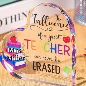 May include: A heart-shaped clear acrylic paperweight with a colorful design that says "The Influence of a great TEACHER can never be ERASED". The design also includes a stack of books, a red apple, and a green sock. The text "Mrs. Miller" is written on the books.