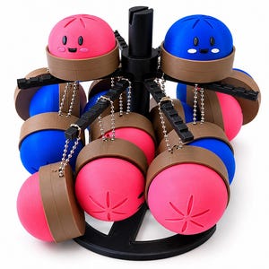 May include: A black rotating display stand holds multiple spherical charms in bright pink and blue. Each charm features a brown base, a smiling face with black eyes, and a star-like pattern on top. The charms are attached to the stand by silver chains.