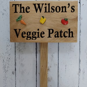 May include: Wooden sign with the text "The Wilson's Veggie Patch" in black lettering. The sign has a brown background and three small plastic vegetables attached to it: a carrot, a bell pepper, and a tomato.
