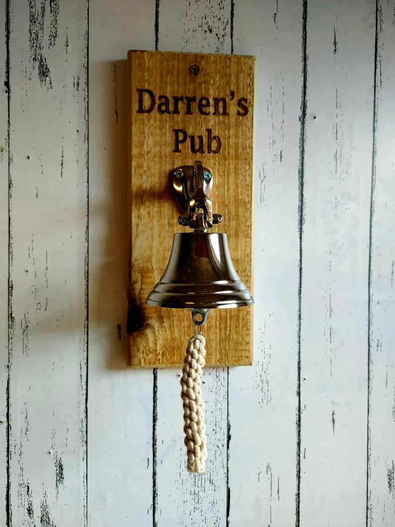 May include: A wooden sign with the text "Darren's Pub" engraved on it. A silver bell with a white rope hangs from the sign.