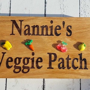 May include: Wooden sign with the text "Nannie's Veggie Patch" in black lettering. The sign has five small plastic vegetables attached to it: a yellow pepper, a carrot, two red cherries, and a yellow lemon.