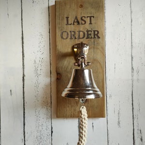 May include: A wooden sign with the text "LAST ORDER" hanging on a white wood wall. A silver bell with a white rope hangs from the sign.