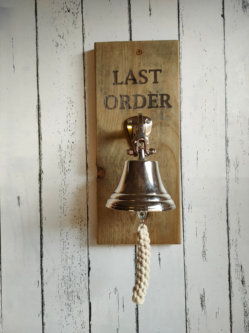 May include: A wooden sign with the text "LAST ORDER" hanging on a white wood wall. A silver bell with a white rope hangs from the sign.