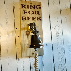 May include: A wooden sign with the text "Ring for Beer" attached to a silver bell with a white rope pull.