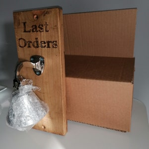 May include: A wooden sign with the text "Last Orders" engraved on it. A silver bell is attached to the sign with a metal hook.