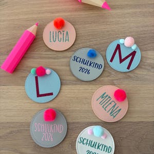 Può includere: Toppe rotonde in tessuto con nomi ricamati e la scritta "SCHULKIND 2026". Ogni toppa ha un colore diverso e un piccolo pompon. Sono presenti anche due grandi matite rosa.