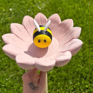 Könnte beinhalten: Ein skurriles Gartenornament mit einer rosa Blume und einer gelb-schwarzen Biene. Die Biene hat weiße Fühler und ein lächelndes Gesicht. Die Blume wird von einem Holzstab gehalten, ideal für den Garten.