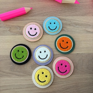 Op de afbeelding: Zeven ronde patches met gestructureerde, pluizige smiley-gezichten in roze, lichtblauw, groen, wit, oranje, geel en felroze. Elke patch heeft een geborduurde zwarte glimlach en is bevestigd op een ronde, gekleurde basis met een gouden rand.