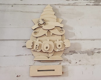 May include: Wooden cutout of a gnome holding pumpkins that spell "BOO". The gnome has a hat, a beard, and a spider on its hat.