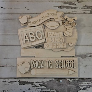May include: A light-colored wooden wall sign featuring school-themed cutouts. The top section displays a banner reading Read-Learn-Grow, an ABC chalkboard, and an apple with a worm wearing glasses. The bottom section features the text Back to School, an apple, and a pencil.