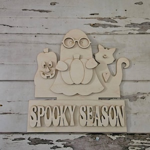 May include: Wooden Halloween decoration featuring a ghost wearing glasses and holding a pumpkin, a jack-o'-lantern, and a cat. The words "Spooky Season" are displayed below the figures. The decoration is made of unfinished wood.