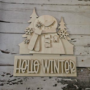 Puede incluir: Decoración de madera de un muñeco de nieve con el texto "HELLO WINTER". El muñeco de nieve tiene una bufanda, botones y una estrella. Dos árboles y copos de nieve también forman parte del diseño. Hecho de madera clara.