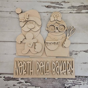 May include: A light-colored wooden cutout sign featuring Santa Claus holding a gingerbread man and Mrs. Claus holding a whisk and mixing bowl. Below the figures, a rectangular wooden plaque displays the text North Pole Bakery in cutout letters.