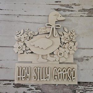 May include: Wooden goose-shaped wall decor with a floral design and the phrase "Hey Silly Goose." The goose has a bow and a tag that says "Honk Honk." The flowers have button centers. The decor is made of light-colored wood.