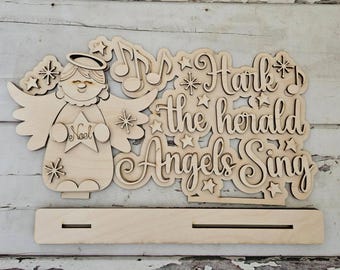 May include: A wooden Christmas decoration featuring an angel holding a star that says "Noel". The words "Hark the herald Angels Sing" are also cut out of the wood, along with musical notes and stars. The decoration has a base for standing.