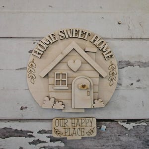 May include: A circular light-colored wooden wall sign featuring the text HOME SWEET HOME, a house illustration with a heart, and a separate rectangular wooden sign below with the text OUR HAPPY PLACE, both with laser-cut details and natural wood grain.