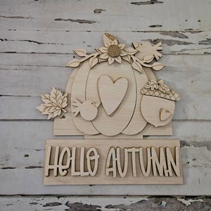 May include: Wooden fall-themed sign featuring a pumpkin, sunflower, birds, and an acorn. The sign includes the words "HELLO AUTUMN" in a decorative font. The sign is made of light-colored wood.