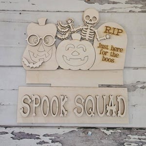 Puede incluir: Un letrero de madera clara para Halloween con una calabaza con gafas, un murciélago, un esqueleto y una lápida con el texto "RIP Just here for the boos". Debajo, una pieza de madera separada muestra el texto "SPOOK SQUAD" en letras grandes.