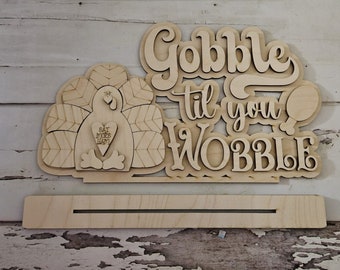 Puede incluir: Un letrero de Acción de Gracias de madera con un recorte de pavo y el texto "Gobble til you wobble". El pavo tiene un corazón con el texto "Eat more ham" en él.