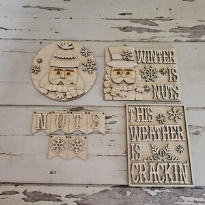 May include: Wooden winter-themed decorations. Includes a round Santa Claus face, a nutcracker, a banner that says "NUTS", and a sign that says "THIS WEATHER IS CRACKIN". All pieces are light brown and feature snowflake accents.